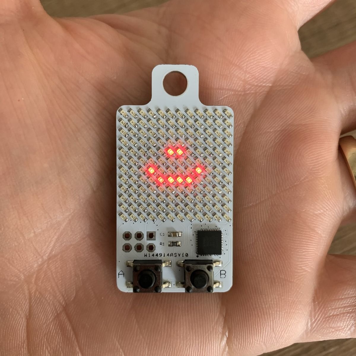 A KeyChainino 144 in the palm of a hand, with a red LED smile lit on its 12 by 12 LED matrix